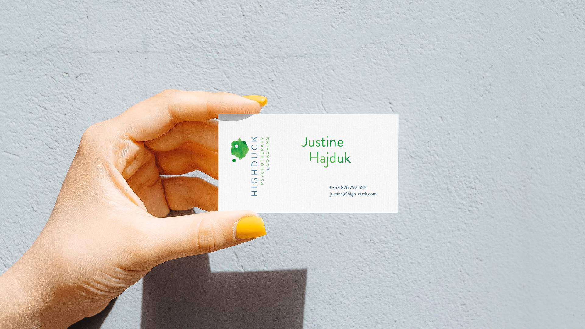 Highduck identyfikacja wizualna - ulotka trzymana w palcach z żółtymi paznokciami, na tle fasady; zdjęcie zrobione w słońcu ponieważ widać cień oraz słoneczne światło