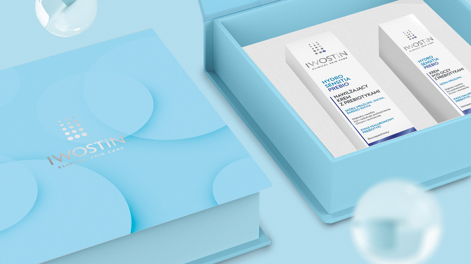 Iwostin opakowanie prezentowe - rzut po skosie; w lewym dolnym rogu część zamkniętego opakowania Iwostin, widoczne przenikające się okręgi z logo, całość niebieska; obok część otwartego pudełka z pokazanym środkiem - produkty