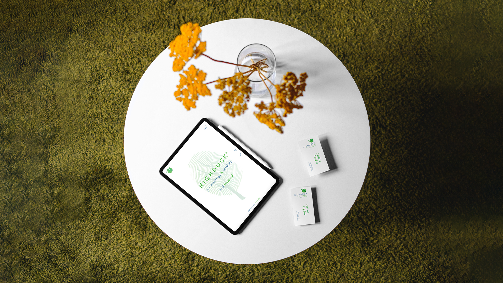Highduck identyfikacja wizualna - biały okrągły stolik w kadrze od góry, umiejscowiony na zielonej ciemnej trawie; na stoliku żółte kwiatki w wazonie i bok rozłożóne dwie wizytówki i tablet z widoczną kolorystyką i elementami identyfikacji HighDuck